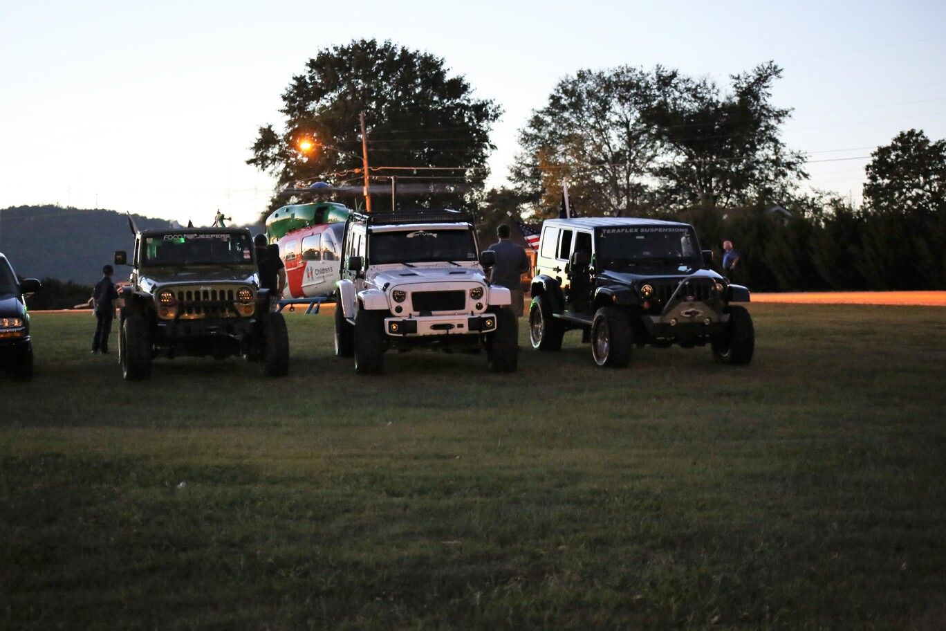 Four off-road Jeeps lined up on a grassy field at dusk, with trees silhouetted in the background.