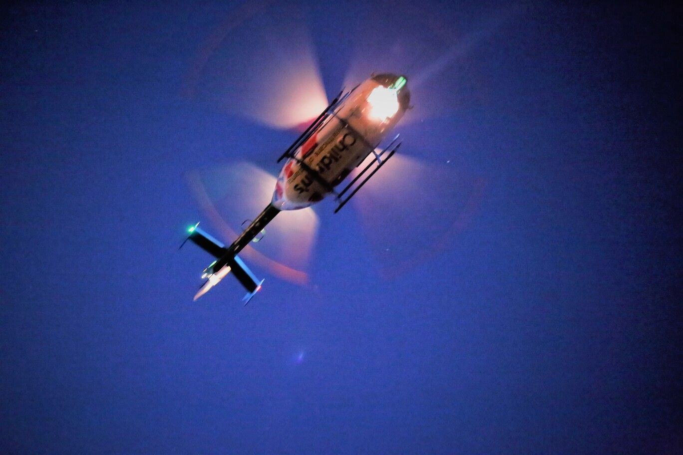 Helicopter flying against a dark blue night sky, with bright rotor lights and spinning blades