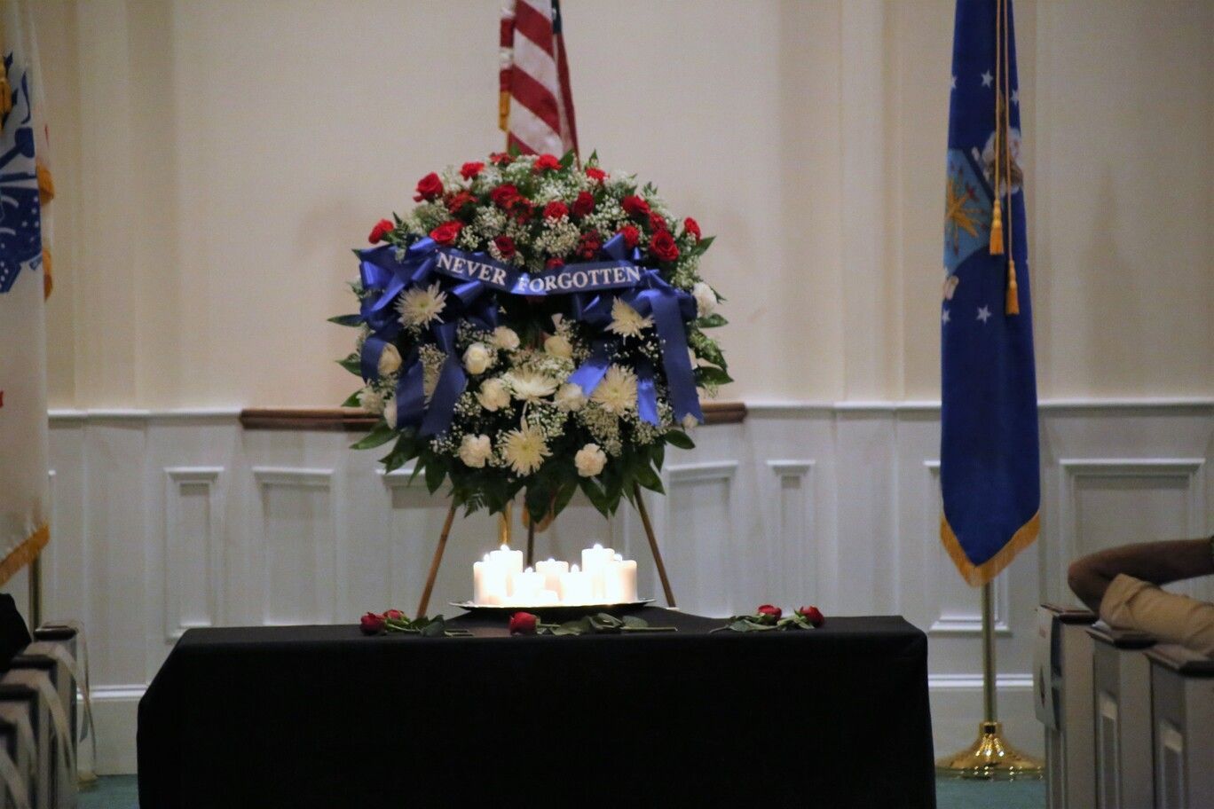 Floral memorial wreath with blue ribbon displayed on a table in a room with flags and chairs.