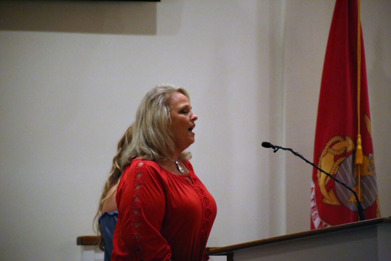 Woman in a red dress speaking at a podium beside a microphone and red flag.