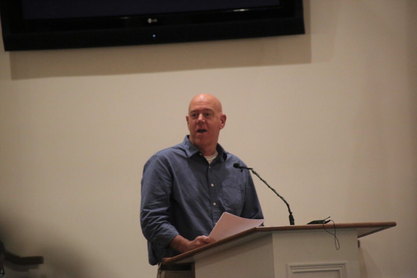 Man speaking at a podium in a conference room, holding papers and facing the audience.
