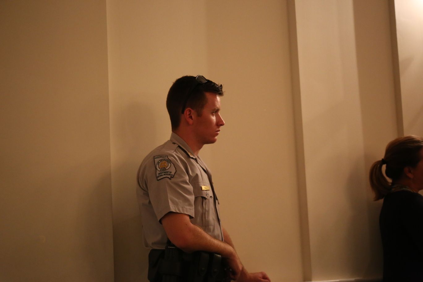 Uniformed officer standing indoors beside a wall, with a person partially visible at right.