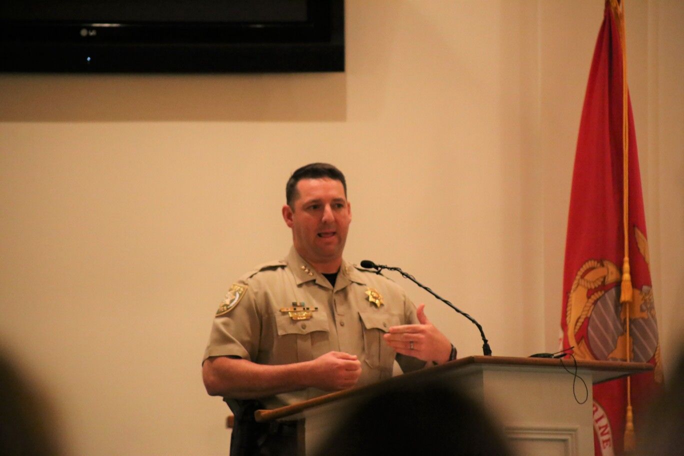 Speaker in tan uniform stands at a podium beside a red flag in a conference room