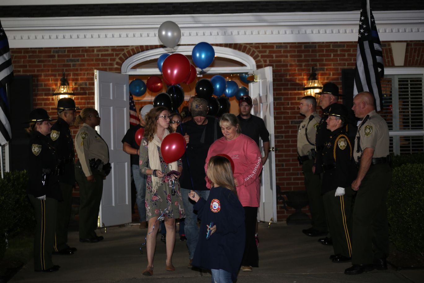 People entering a brick building at night under balloon arches, surrounded by uniformed officers.