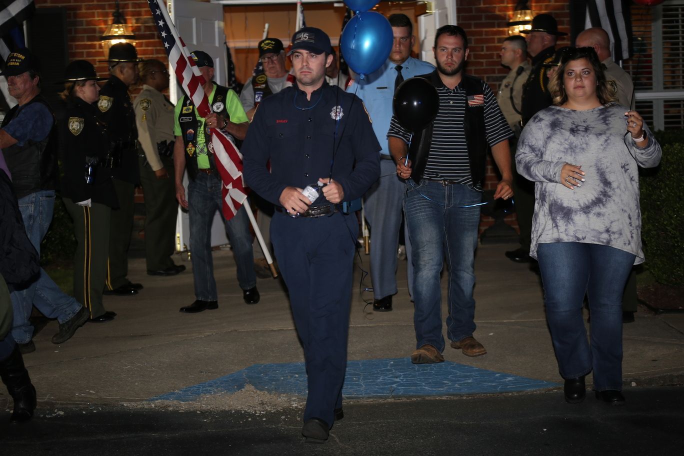 Uniformed officer leads a small crowd through a dimly lit event space with balloons and American flags.