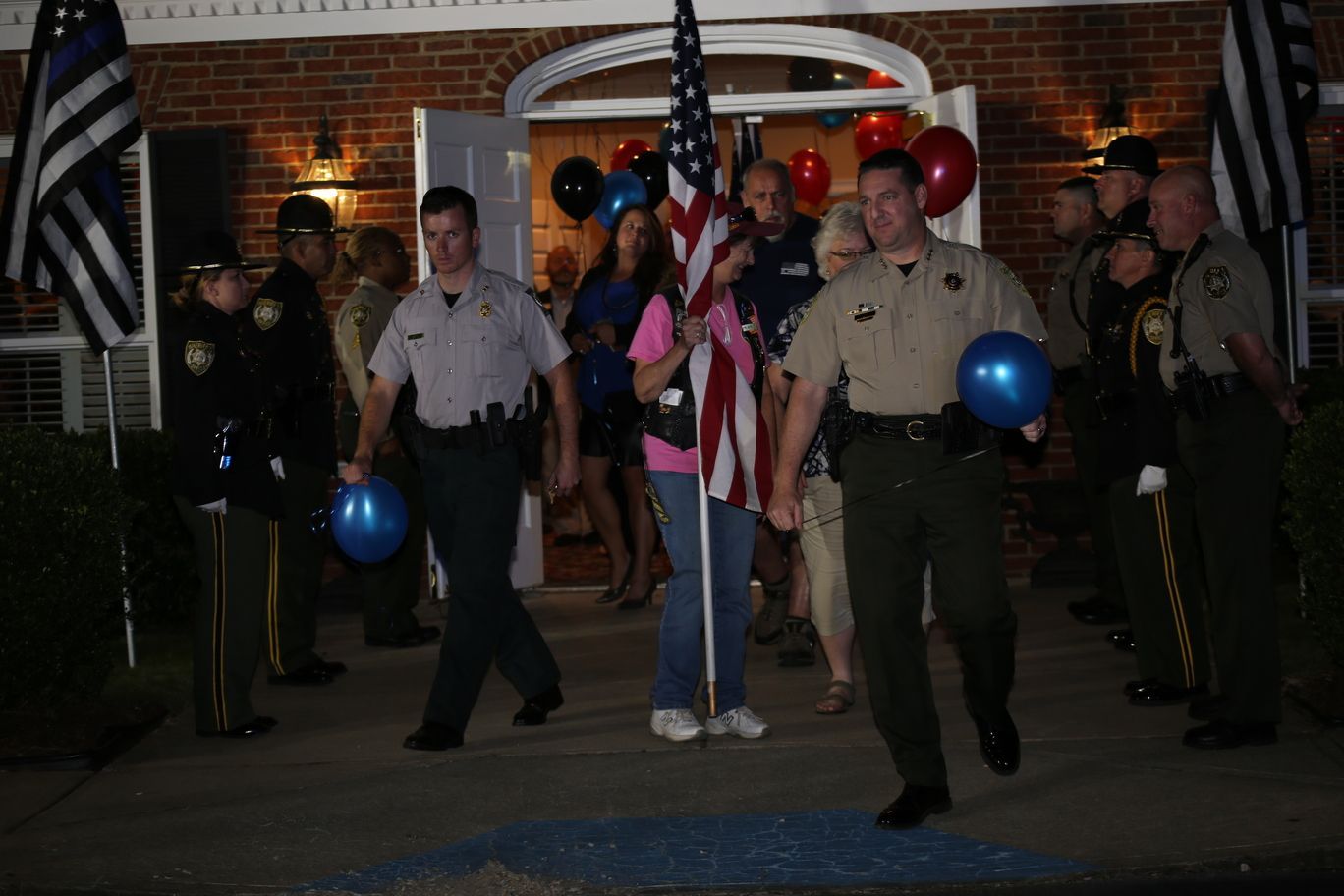 Uniformed officers holding blue balloons lead a nighttime procession with American flags in front of a building.