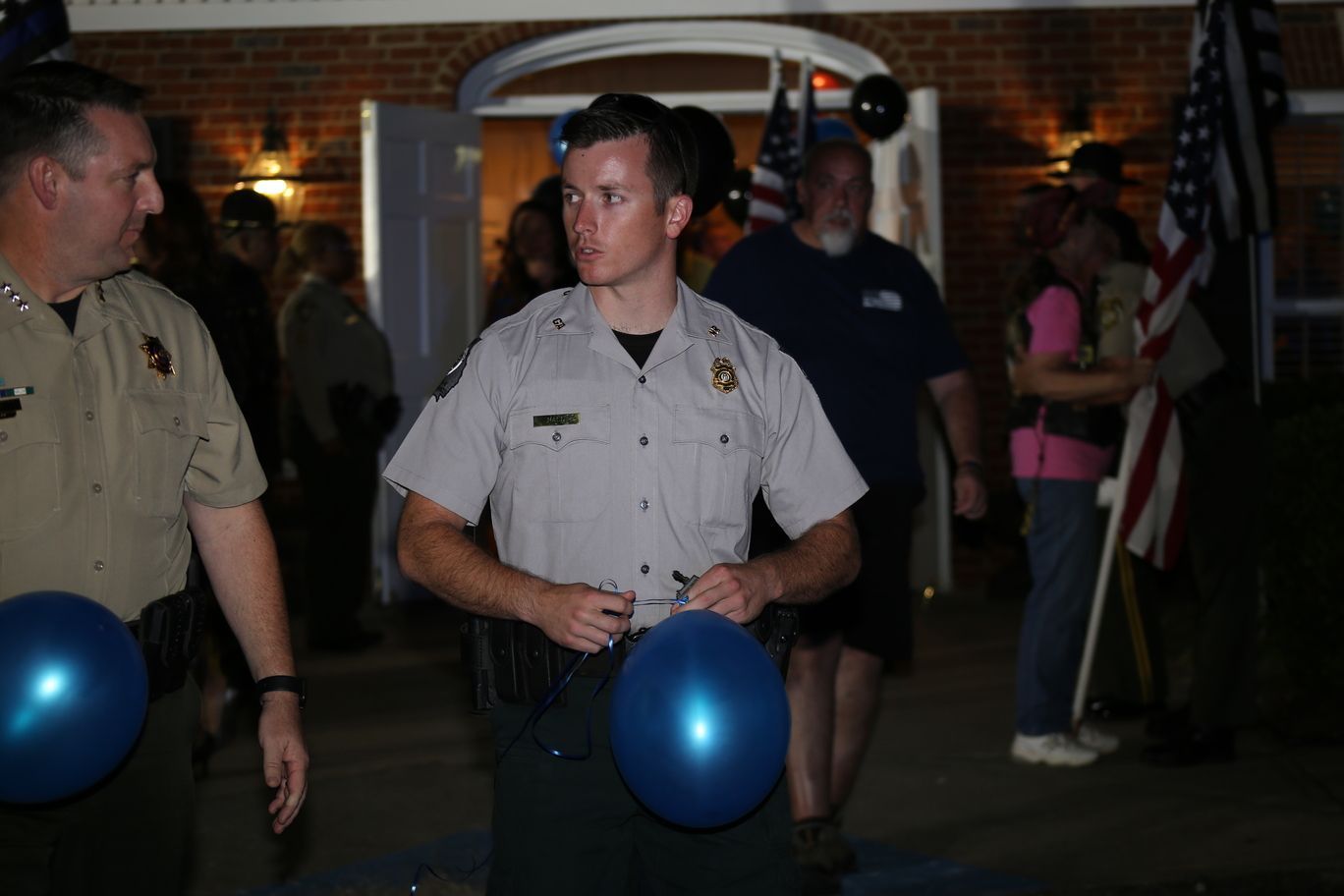People at a nighttime gathering hold blue balloons outside a brick building.