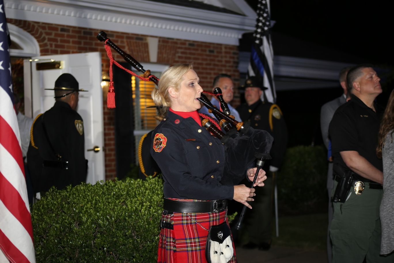 Person playing bagpipes at a nighttime outdoor event, with officers and a house in the background.