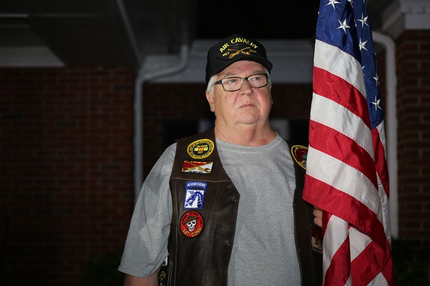 Veteran standing beside an American flag, wearing a cap and vest with military patches.