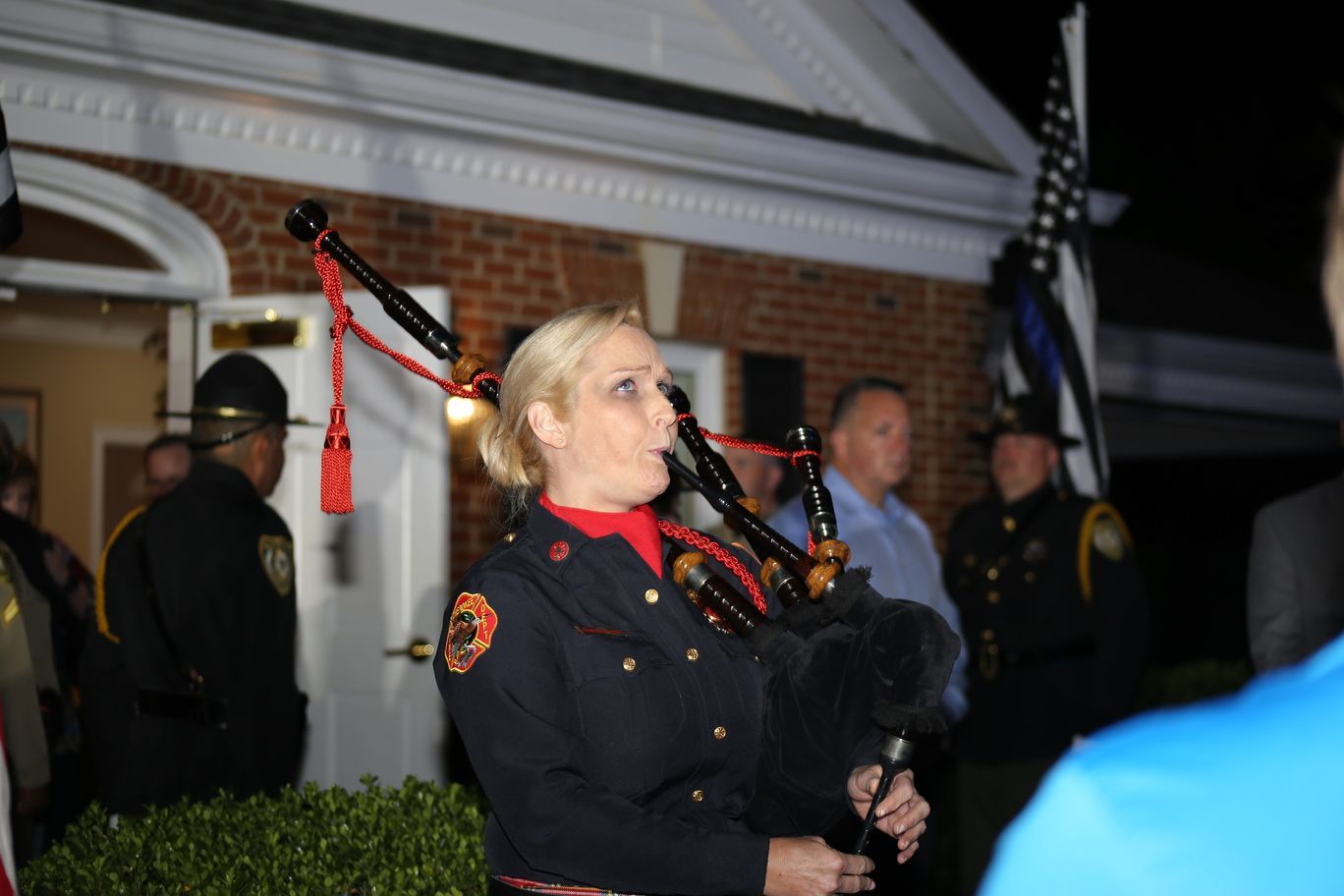 Bagpiper in dark uniform playing outdoors at a nighttime ceremony with police officers nearby
