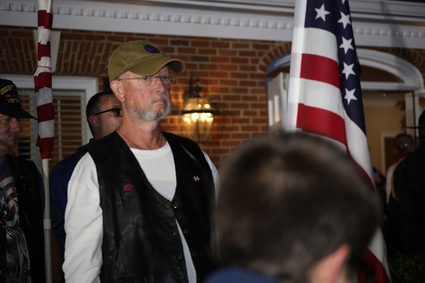 Man in tan cap and black vest stands indoors beside American flags at a brick-walled gathering