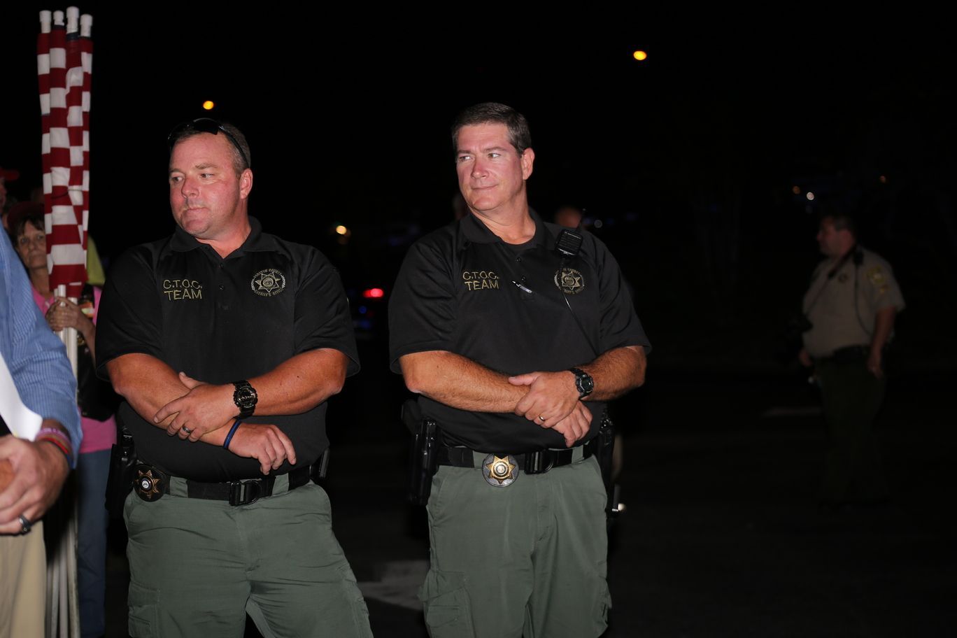 Two uniformed officers stand at night with arms crossed, near an American flag.
