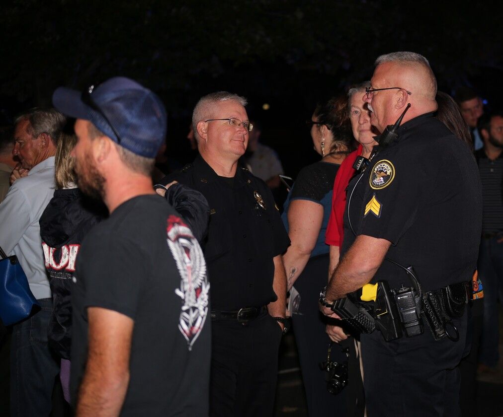 Police officers and civilians standing together at night, talking in a dark outdoor scene