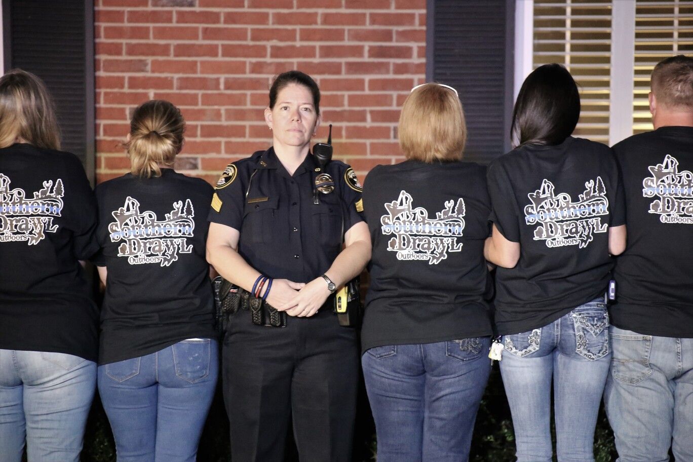 Police officer standing with six people in black shirts, facing a brick wall.