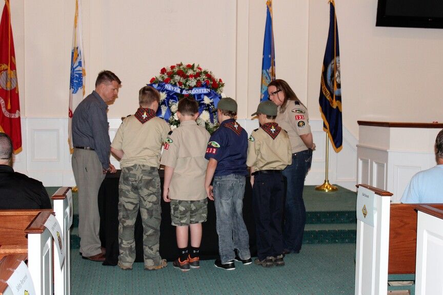 Scouts in tan uniforms stand on a church stage during a ceremony, with flags and floral arrangement behind them.