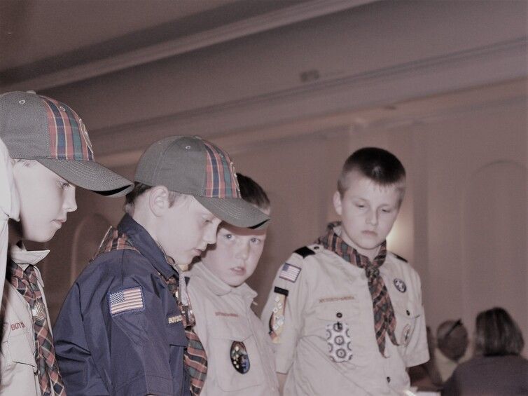 Boy Scouts in uniform standing together indoors, looking down at something off-camera.