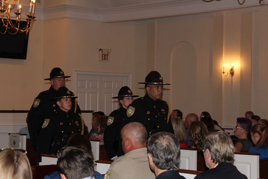 Police officers in dark uniforms and hats stand in a room with seated audience members.