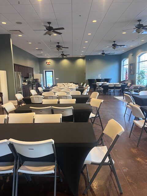 Empty banquet hall with black tablecloths, white folding chairs, ceiling fans, and large windows.