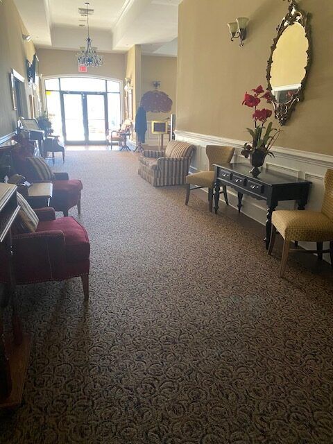 Long carpeted hallway with chairs, tables, mirrors, and sunlight from glass doors at the end.