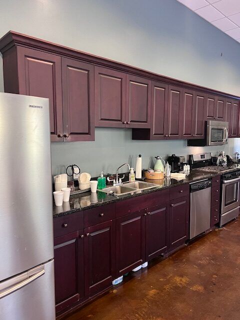 Commercial kitchen with maroon cabinets, stainless fridge, and countertop appliances along a tiled wall.