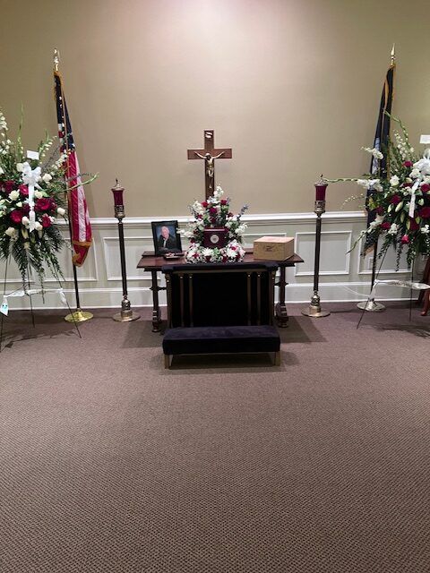 Church altar with cross, floral arrangements, flags, and a book on a table in a carpeted hall