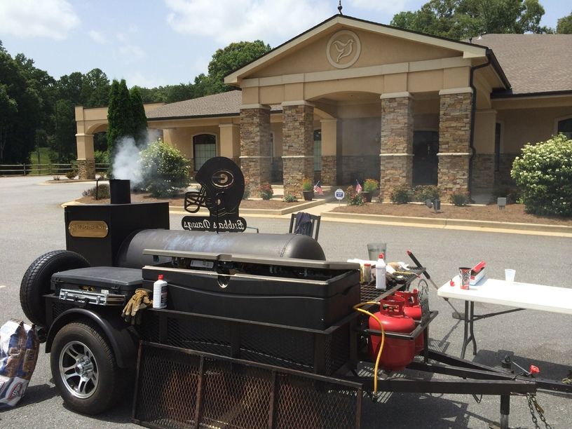 Black barbecue smoker trailer parked outside a stone building, with smoke rising from the cooker.