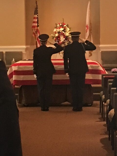 Two uniformed guards saluting beside a flag-draped casket with a flower wreath