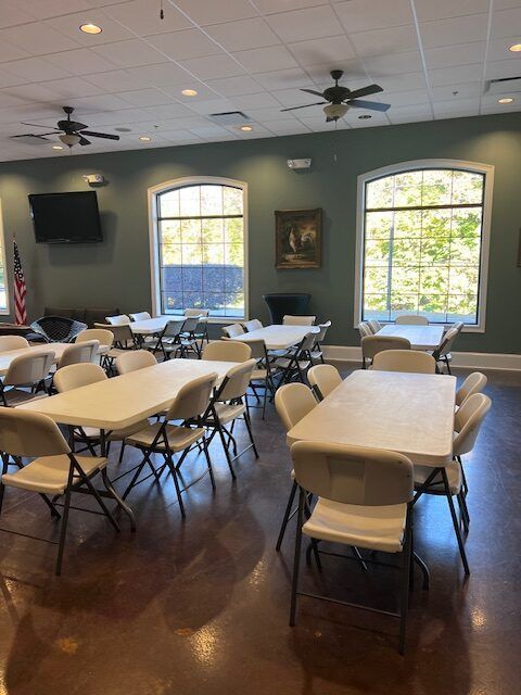 Empty meeting room with tables and chairs, TV screen, ceiling fans, and large windows with daylight.