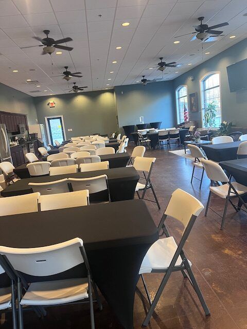 Empty dining hall with black tables, white folding chairs, ceiling fans, and large windows.
