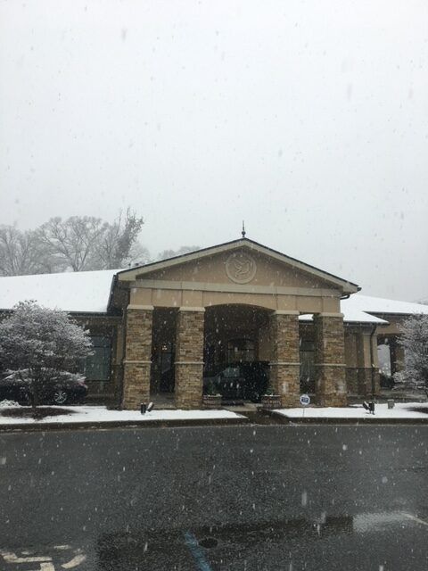Snowy view of a beige building with columns and a dark roof across a wet road