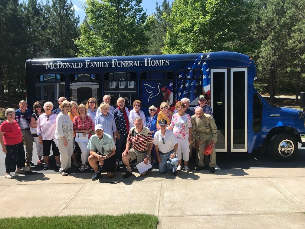 Group posing beside a blue funeral home bus in a sunny outdoor parking lot