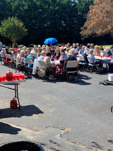People seated at long tables outdoors in bright sunlight, with drinks and folding chairs on a paved lot.