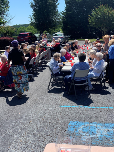 Outdoor community gathering with people seated at folding tables in a sunny parking lot, sharing a meal.