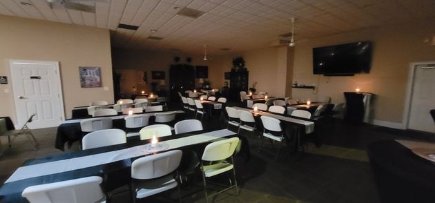 Empty banquet hall with rows of white-covered chairs and tables, wood-paneled walls, and a wall-mounted TV.