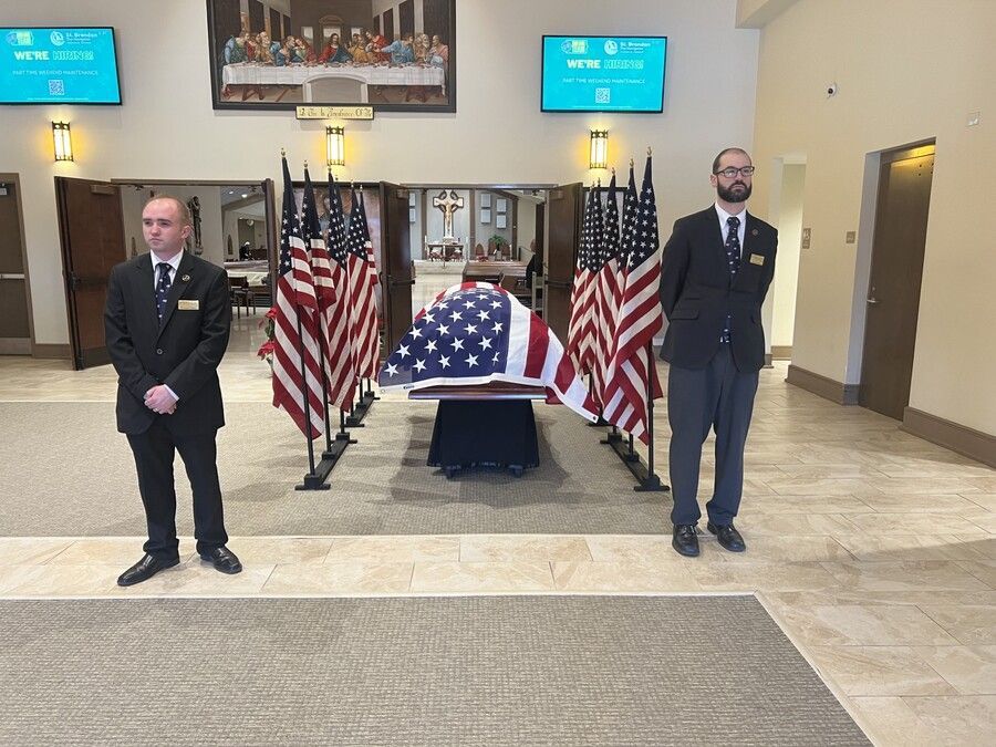 Flag-draped casket in a formal hall, flanked by American flags and two uniformed honor guards.