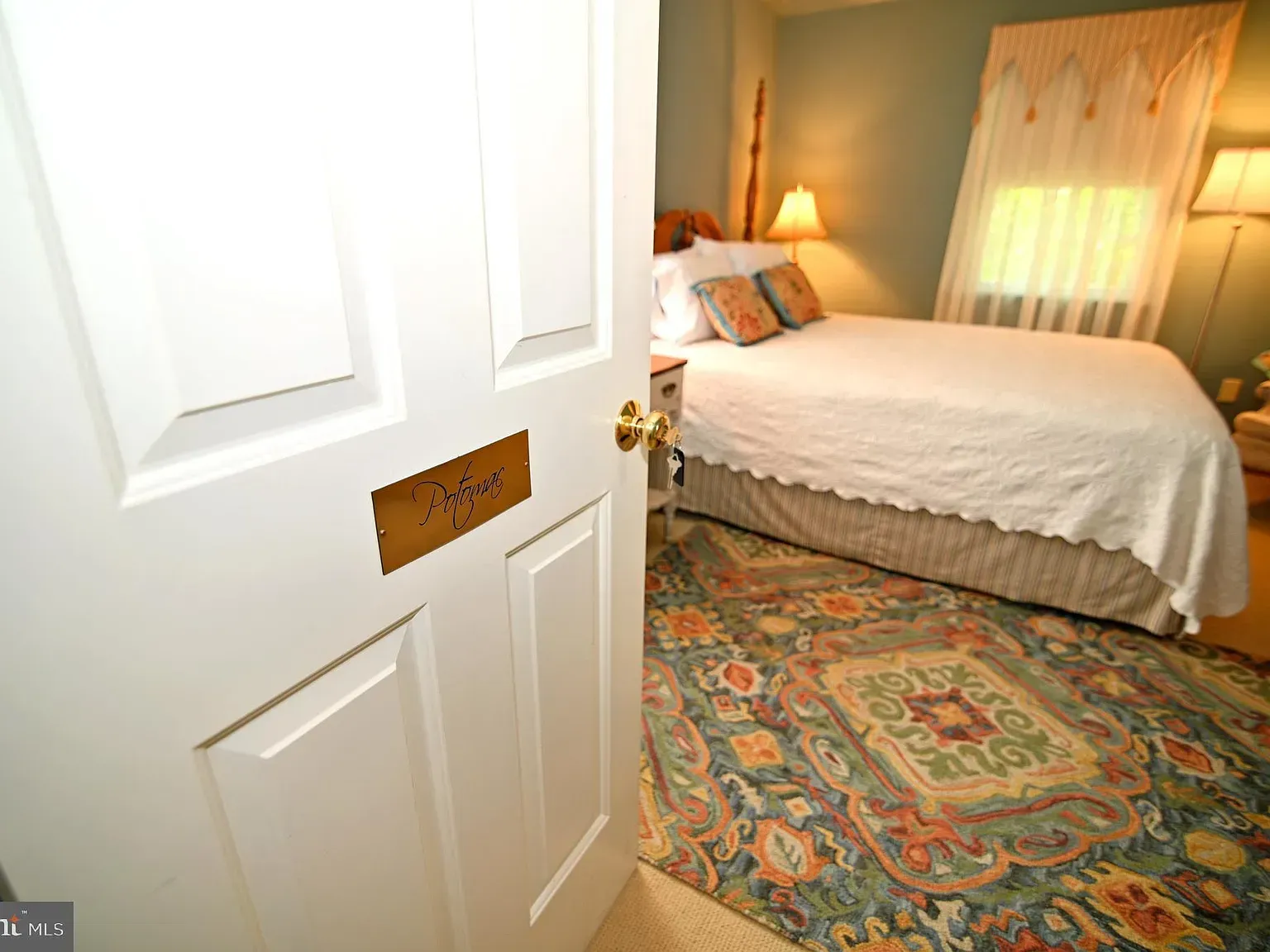 Bedroom interior: white door ajar revealing bed, rug, and window with curtains; light colors dominate.