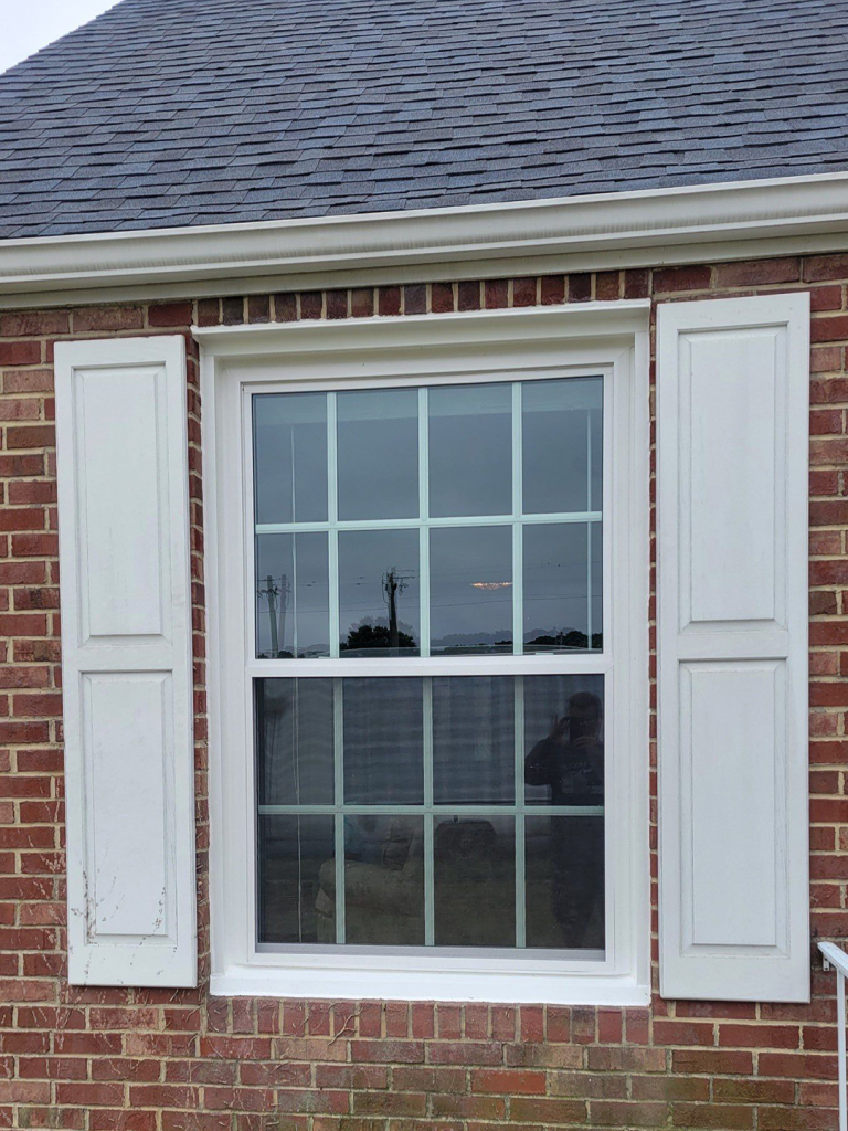 Brick house window with white shutters and a gray shingled roof reflection