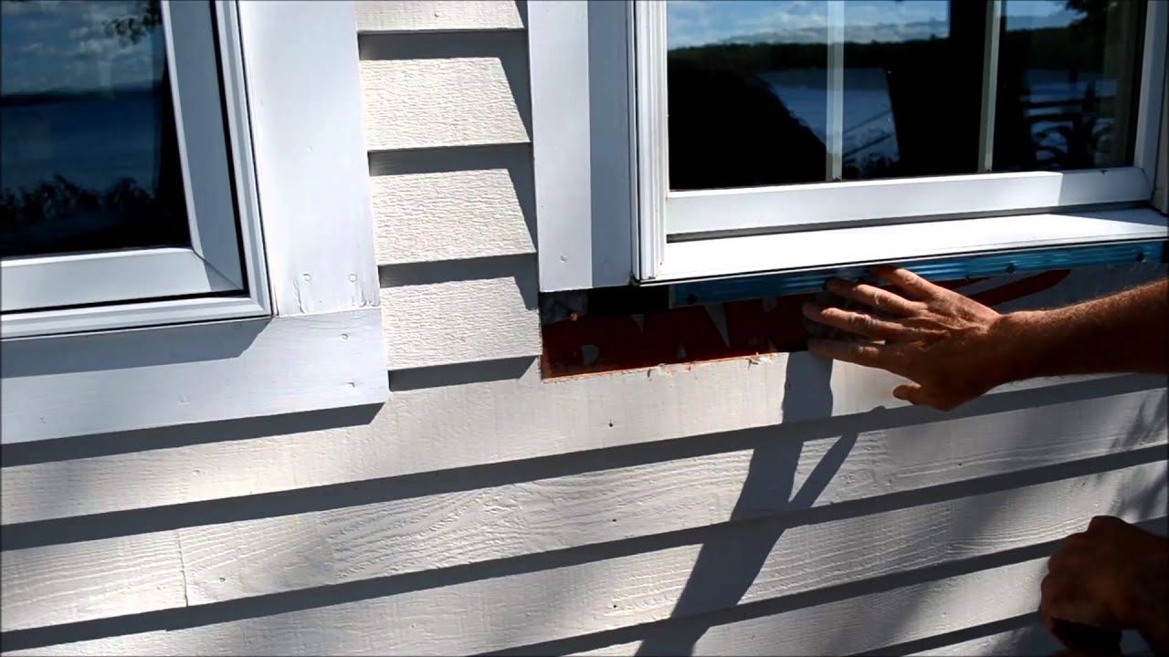 Hand pointing at a red-brown ledge beneath a white window on a light gray house exterior