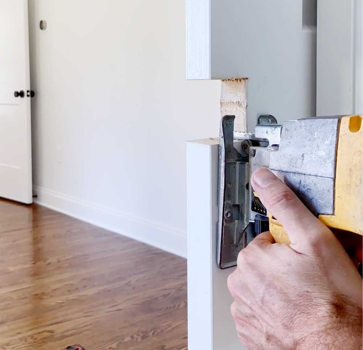 Hand screwing a metal hinge onto a white cabinet door in a bright room with hardwood floors