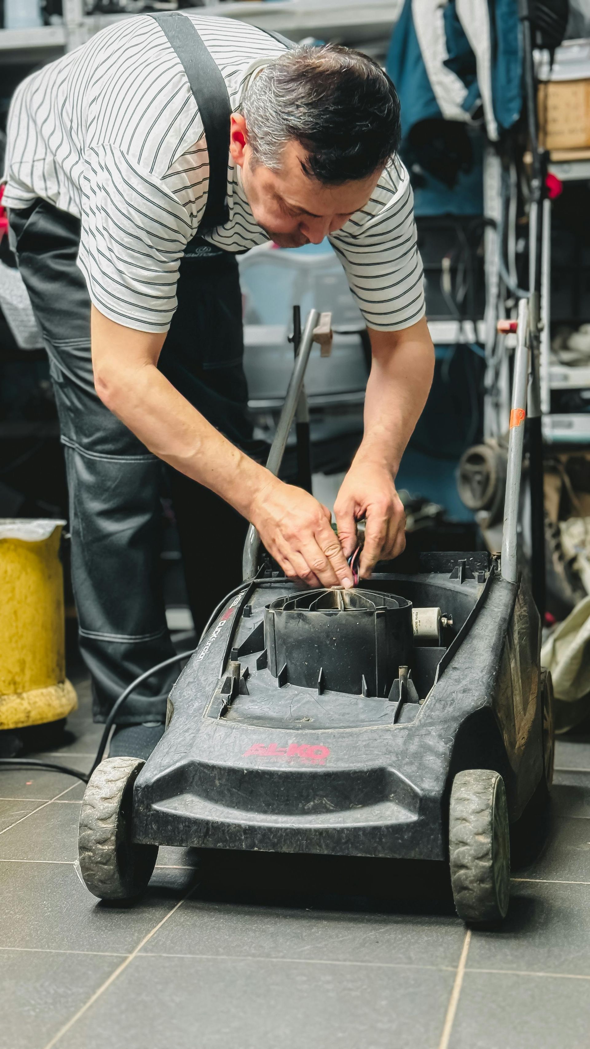 Man in overalls repairing a lawnmower in a cluttered garage.