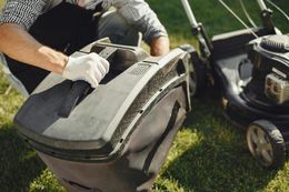 Person in gloves attaching a grass catcher to a black lawnmower on a green lawn.