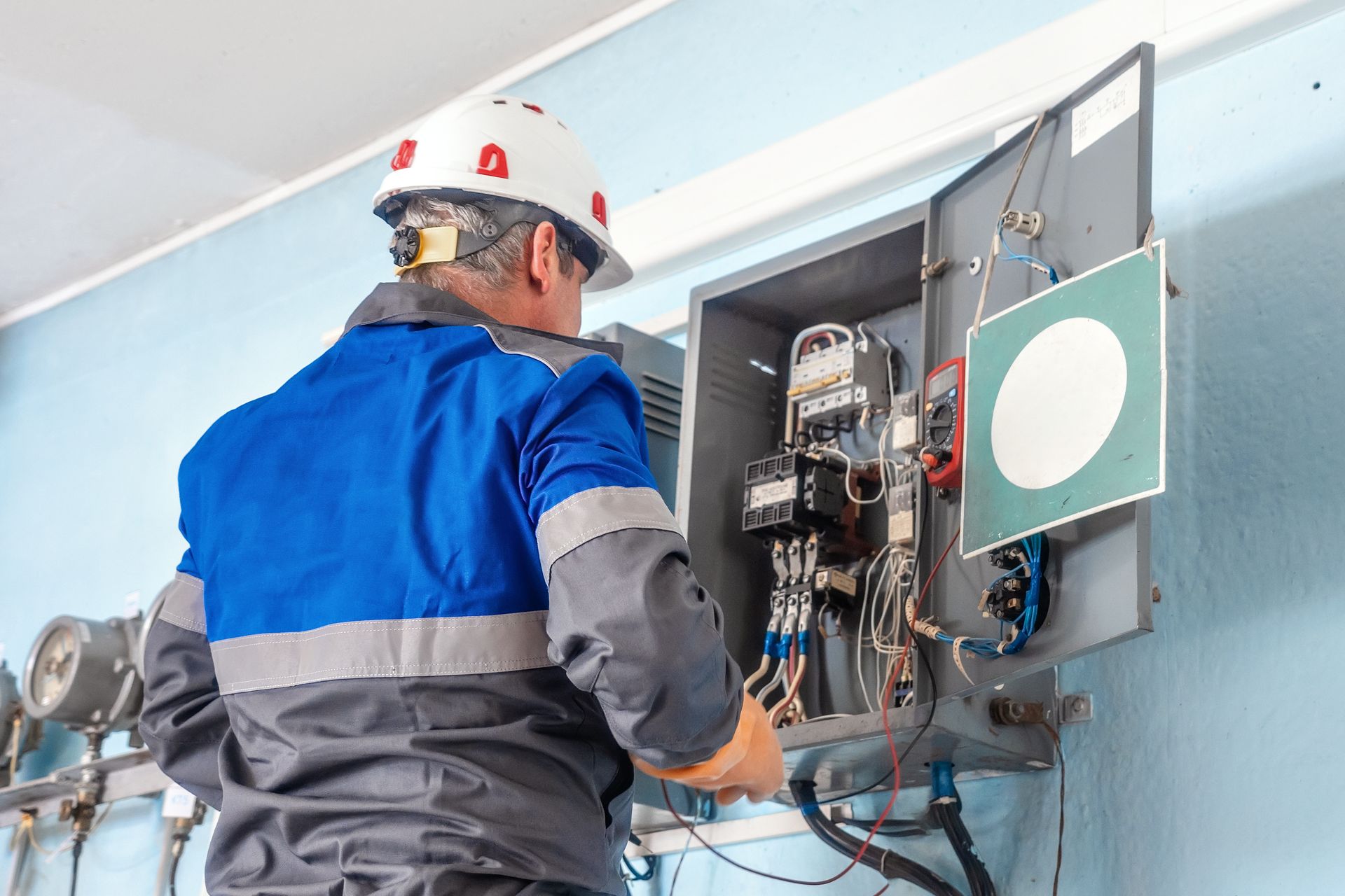An electrician in a hard hat testing wiring inside a commercial electrical panel.