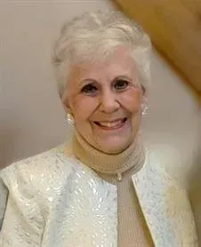 Smiling woman in a white textured jacket and beige turtleneck against a soft indoor background