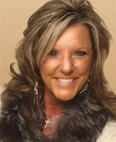 Smiling woman with blonde hair wearing a fur-collared coat and cross necklace against a beige background