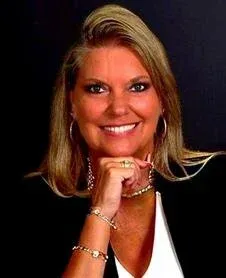 Smiling woman with blonde hair in a black blazer and white top, resting her chin on her hand
