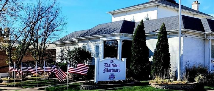 White house with American flags and a sign reading “The Trinity,” set in a sunny landscaped yard