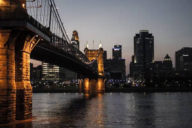 A bridge with lit stone pillars over a dark river at dusk, with city skyline in the background.