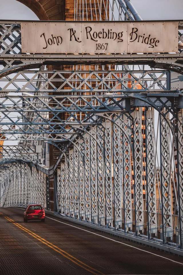 Red car crossing John A. Roebling Bridge. Steel arch with sign