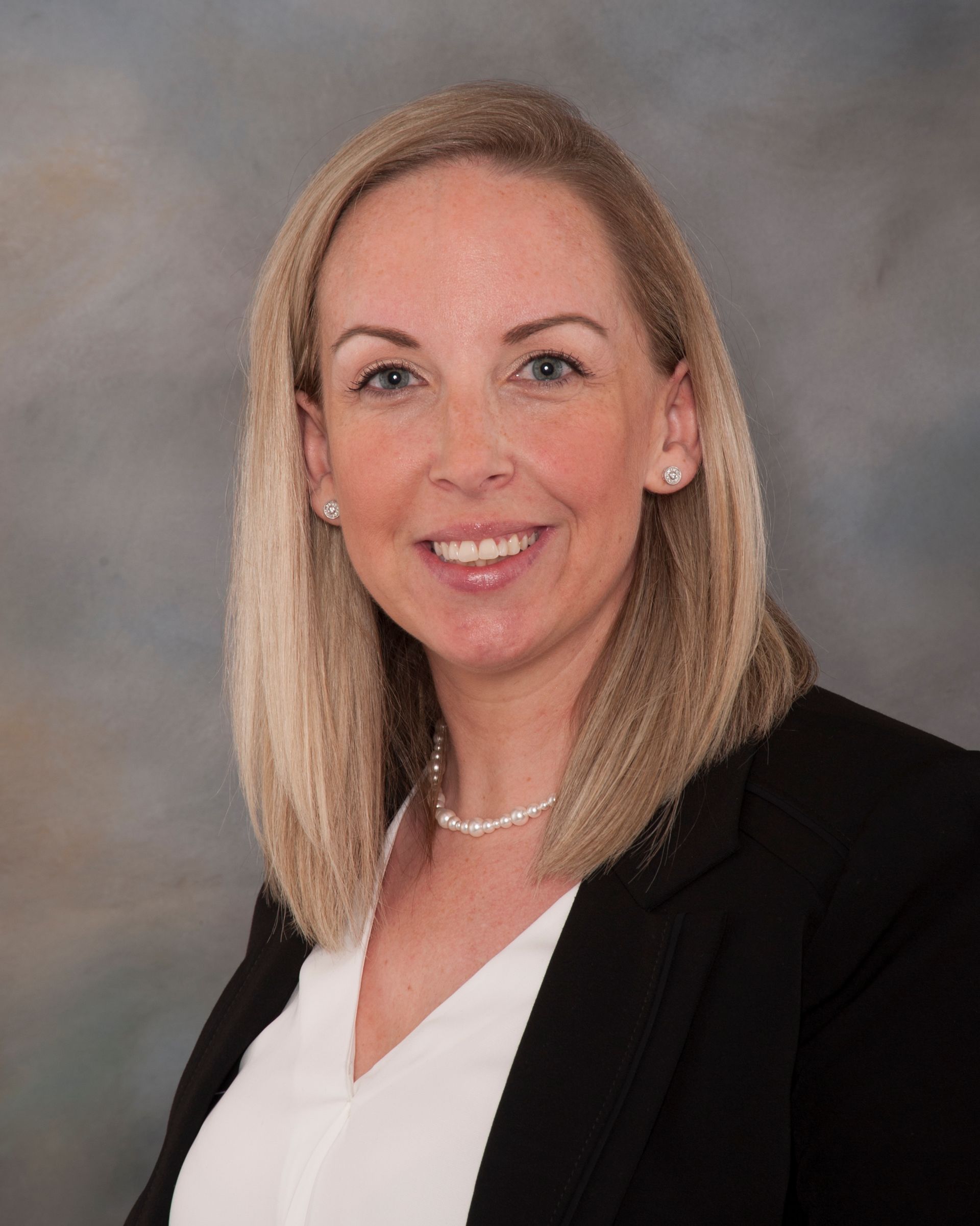 Woman with blonde hair, smiling, wearing a white shirt, black blazer, and pearl necklace.