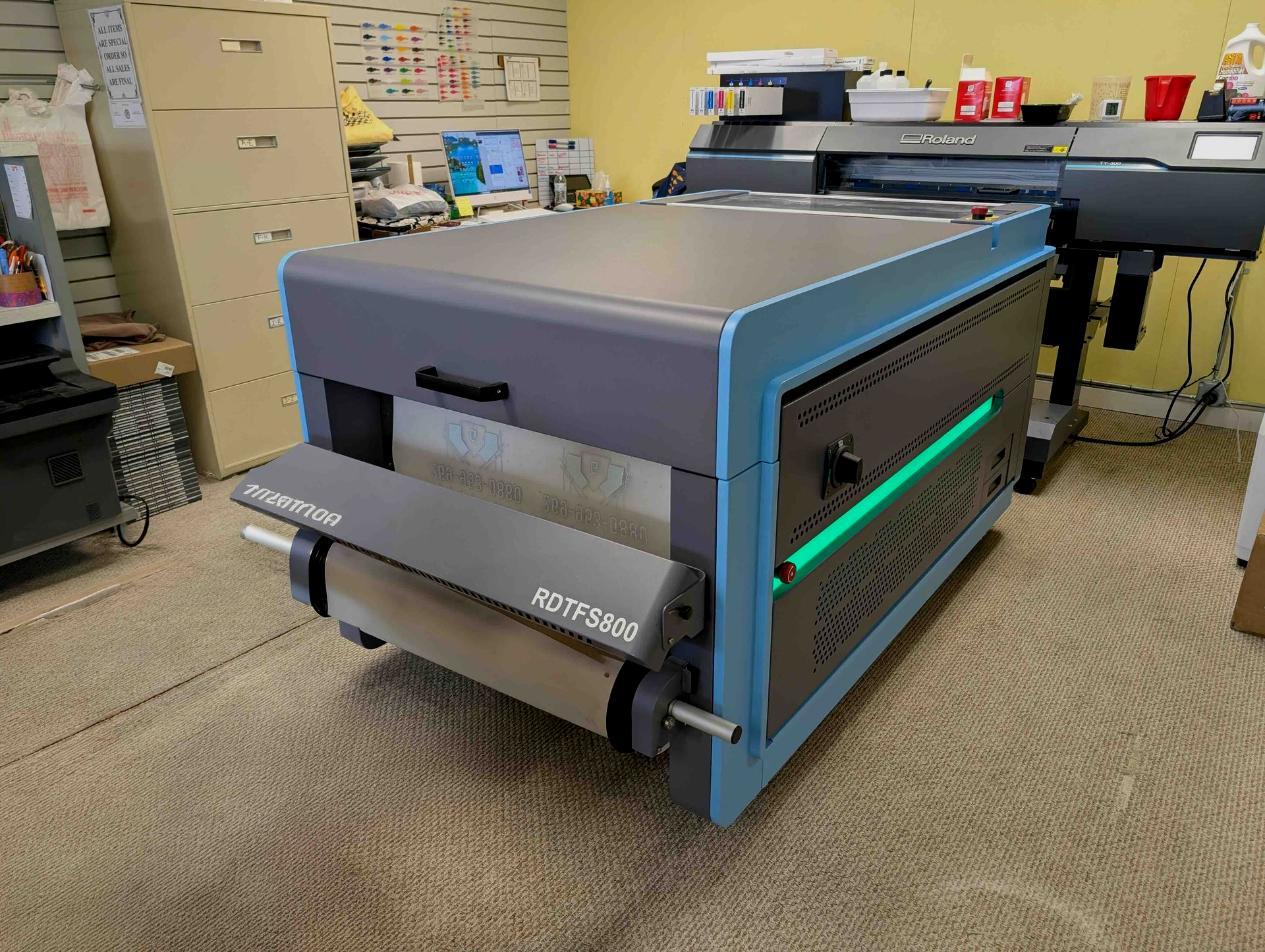 Large blue industrial printer in a workshop, with paper rolls and computer equipment nearby.
